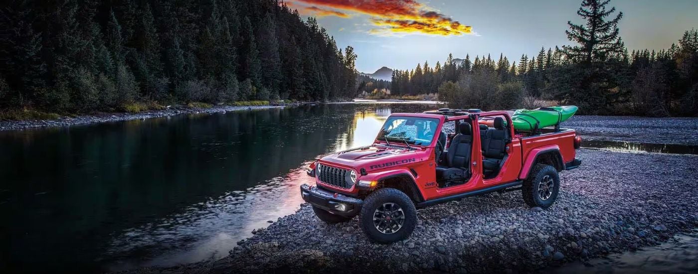 A red 2024 Jeep Gladiator Rubicon with a mounted kayak parked by a river.